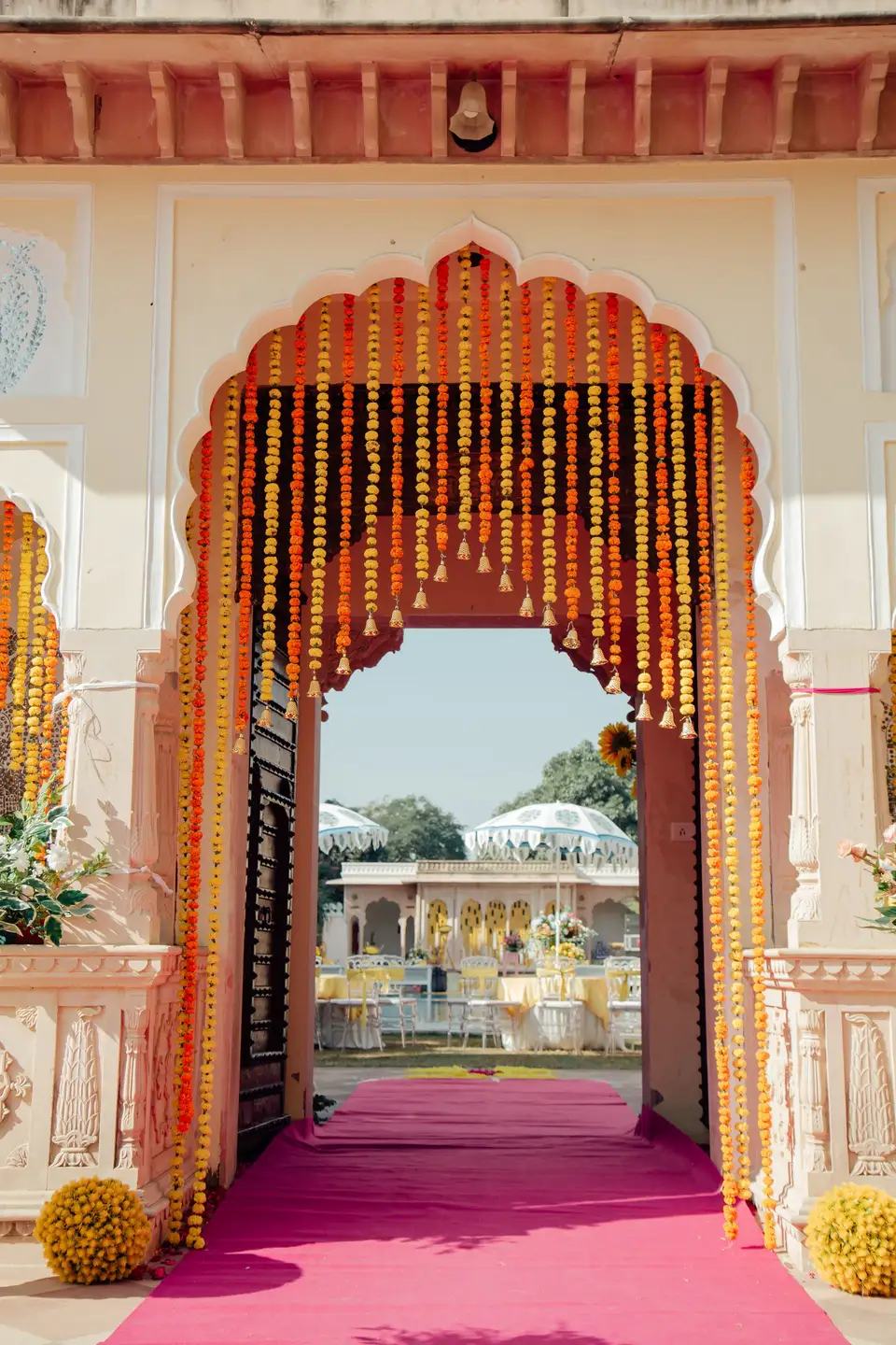 A wedding entrance, an ornate scalloped archway hung with a dense curtain of yellow and orange marigold garlands, a pink carpet running through it into a sunlit courtyard beyond.