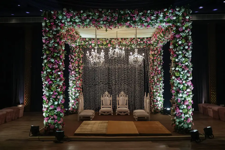 A dressed indoor wedding mandap, a floral arch in pink and white around a sequinned backdrop, two carved thrones on a low stage and three crystal chandeliers hanging above.